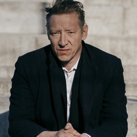Billede af Christian Groes. Sidder i sort jakke med murstensbaggrund